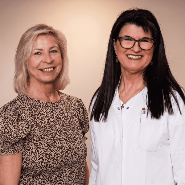 Ein lächelndes Portraitbild von Carmen und Dr. med. Sabine Ott-Oechsle, die nebeneinander vor einem hellen Studiohintergrund stehen. Carmen trägt eine Bluse im Leopardenmuster und Sabine Ott-Oechsle einen weißen Arztkittel und eine Brille.